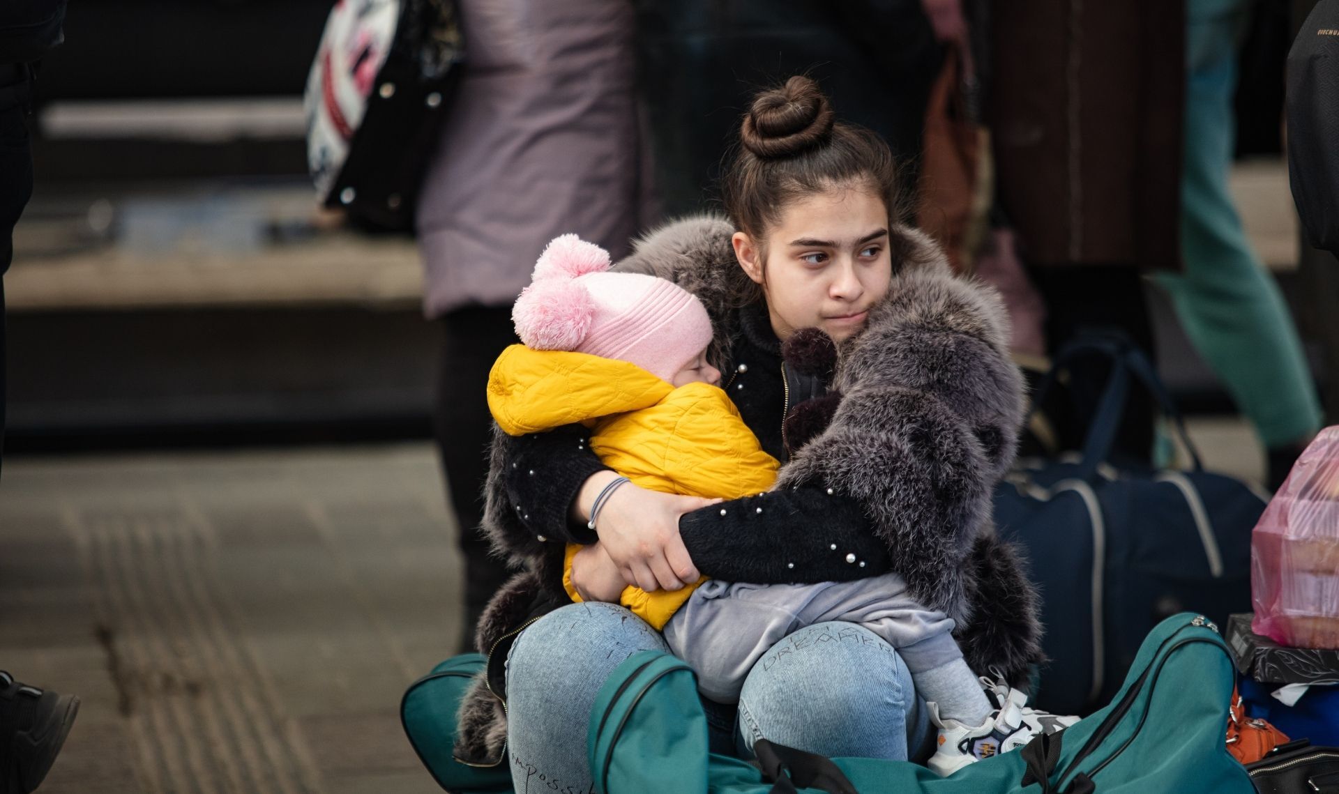 Mother holds her child, wrapped in warm clothes with packed bags beside them